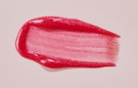 Close-up of a glittery red lip tint smeared in a swipe on a pink background, showing its glossy, sparkly texture