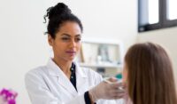 A clinician wearing a white coat and gloves examines a patient’s face during a consultation.